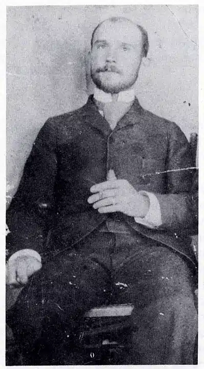 A grainy black and white photo of a young man in a three-piece suit, seated and looking at the camera.